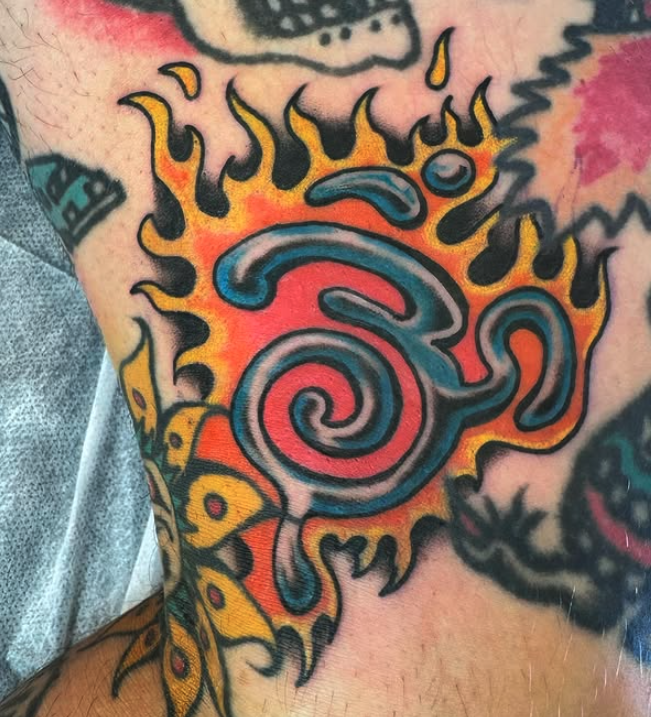 Colorful tattoo of a spiral on skin with flames in yellow, orange, and black surrounding it.