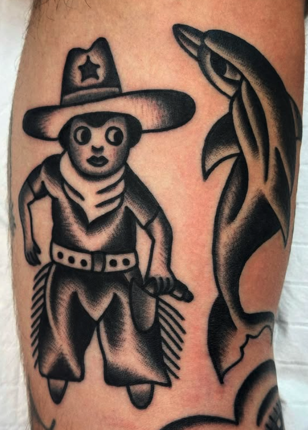 A black and gray tattoo of a cartoon cowboy with a large hat, scarf, belt, and boots, holding a small object, situated next to a large fish on a person's skin.