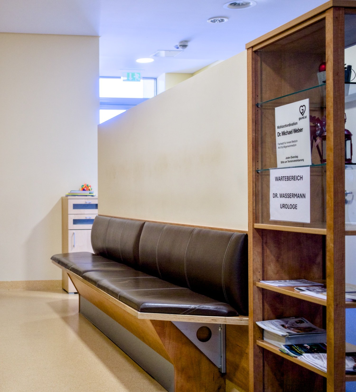 Wartebereich in einer medizinischen Einrichtung mit braunem Sitzbank, Holzregal mit Informationsmaterial und Hinweisen auf Wahlorganisation, Dr. Michael Weber, Dr. Wassermann. Helle Wände, Fenster und Deckenleuchten im Hintergrund.