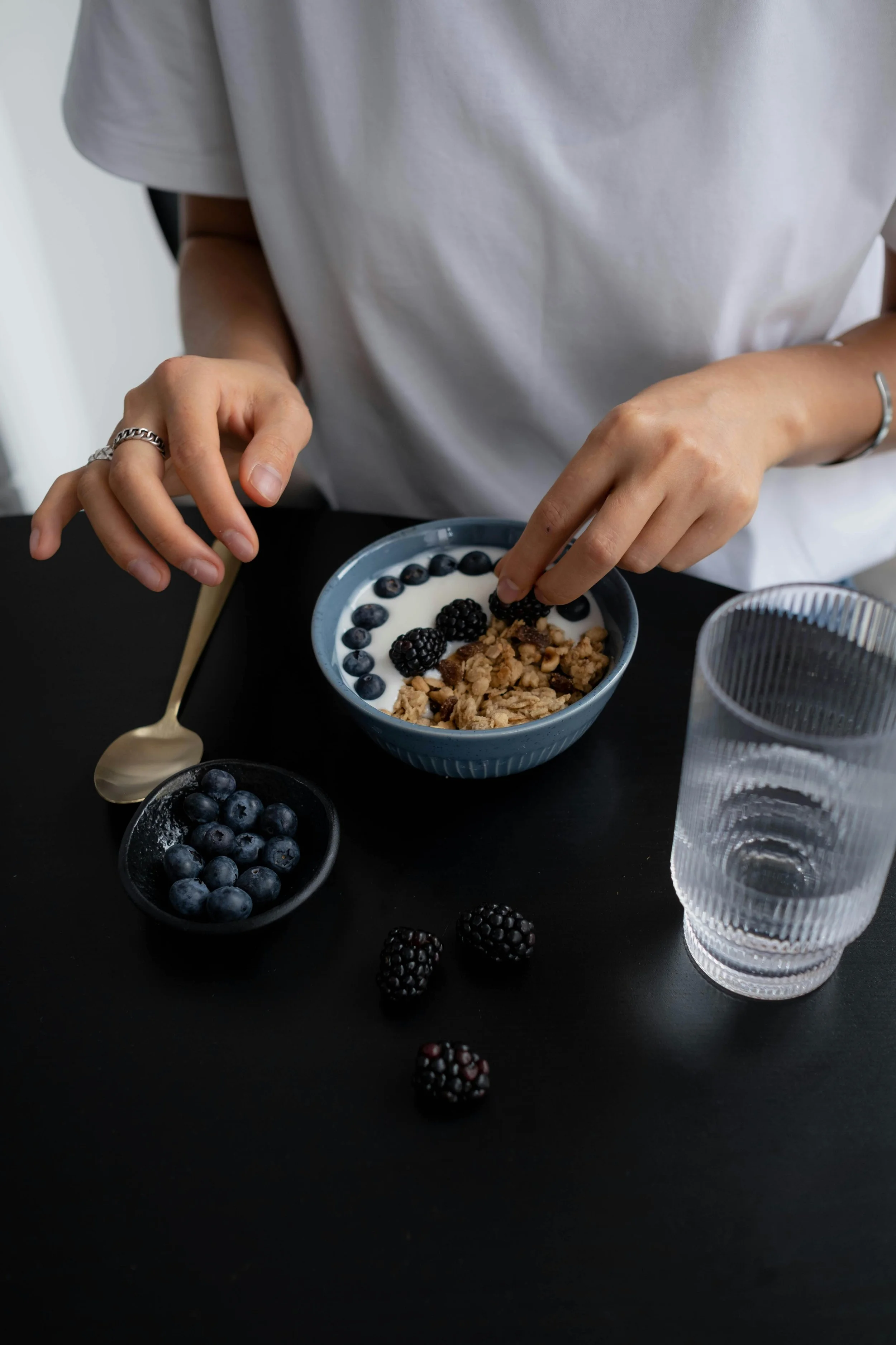 A person in a white shirt preparing a bowl of yogurt topped with blackberries, blueberries, and granola, with a small bowl of blueberries, a glass of water, and a gold-colored spoon on a black table.