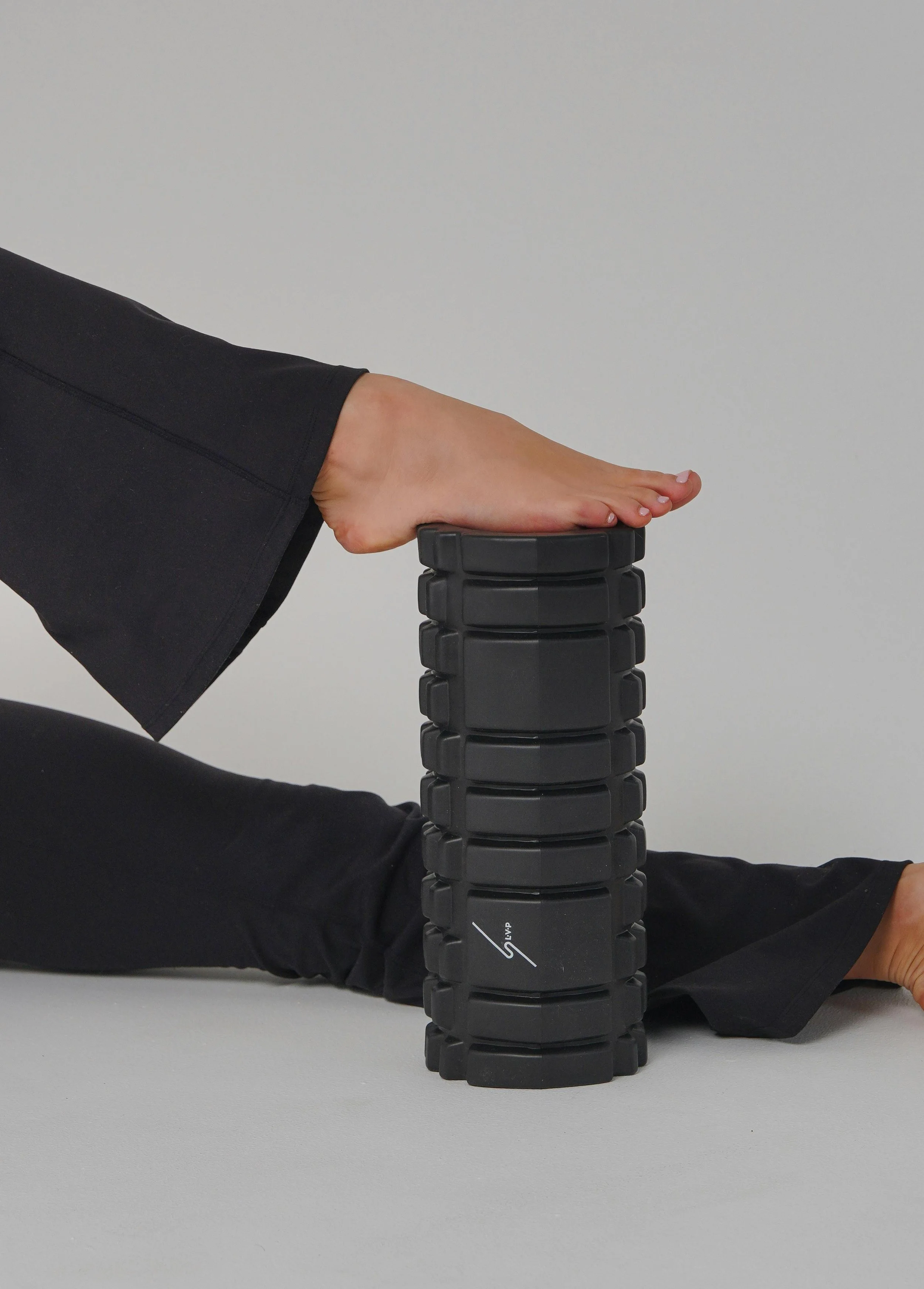 Person with black pants practicing yoga or stretching on a gray surface, placing their hand on a black, segmented yoga or massage roller with multiple joints.