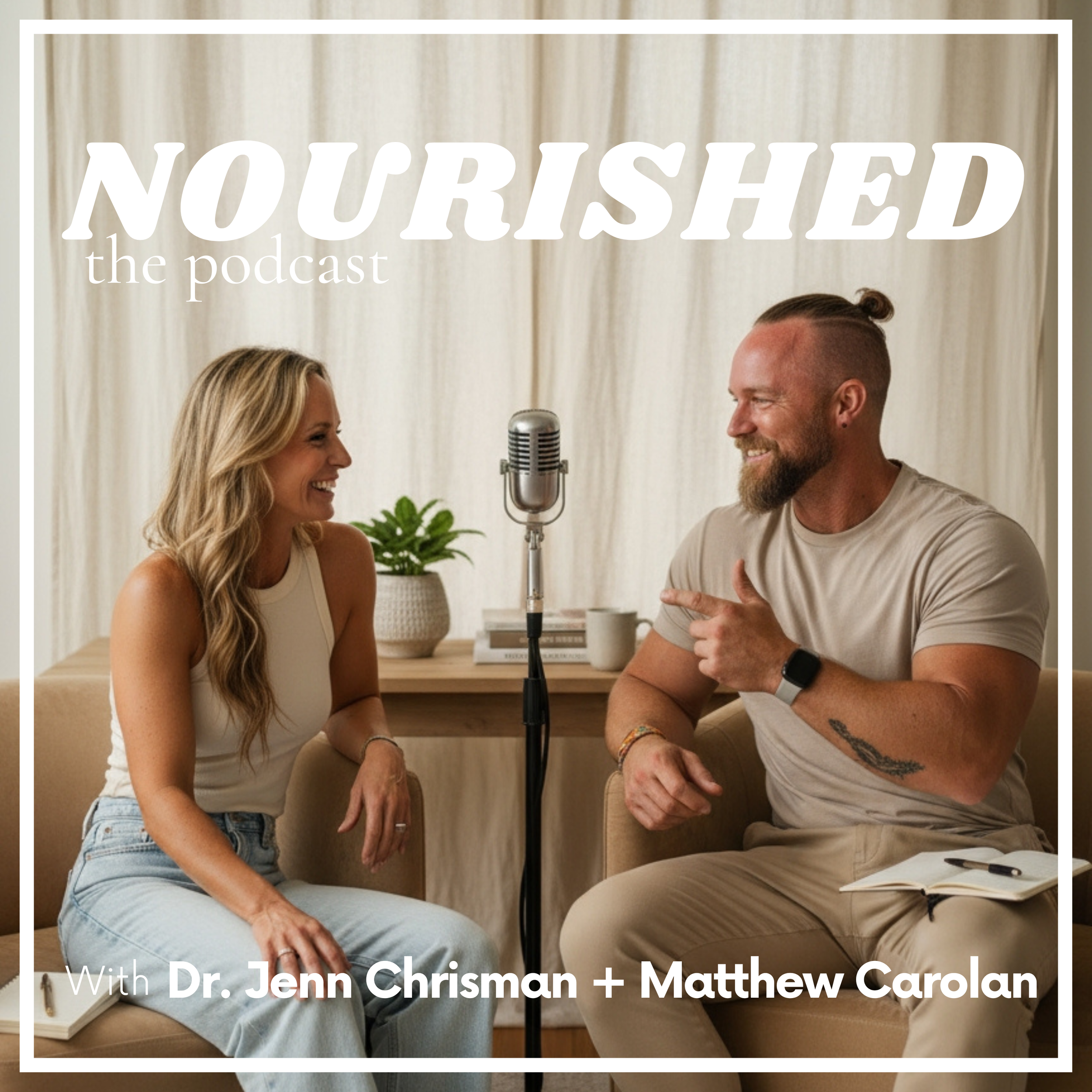 Two people, a woman and a man, sitting and talking with a microphone between them. The woman has long blonde hair and is wearing a sleeveless top, and the man has a top knot and beard, and is wearing a t-shirt and a smartwatch. They are smiling and engaging in conversation in a cozy, well-lit room.