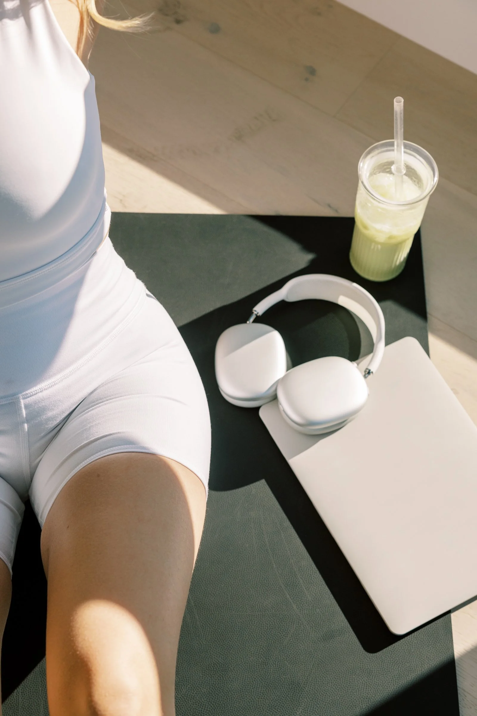 A person in white workout clothes sitting on a black yoga mat with white headphones, a closed white laptop, a green smoothie with a straw, and a white notebook on the mat.