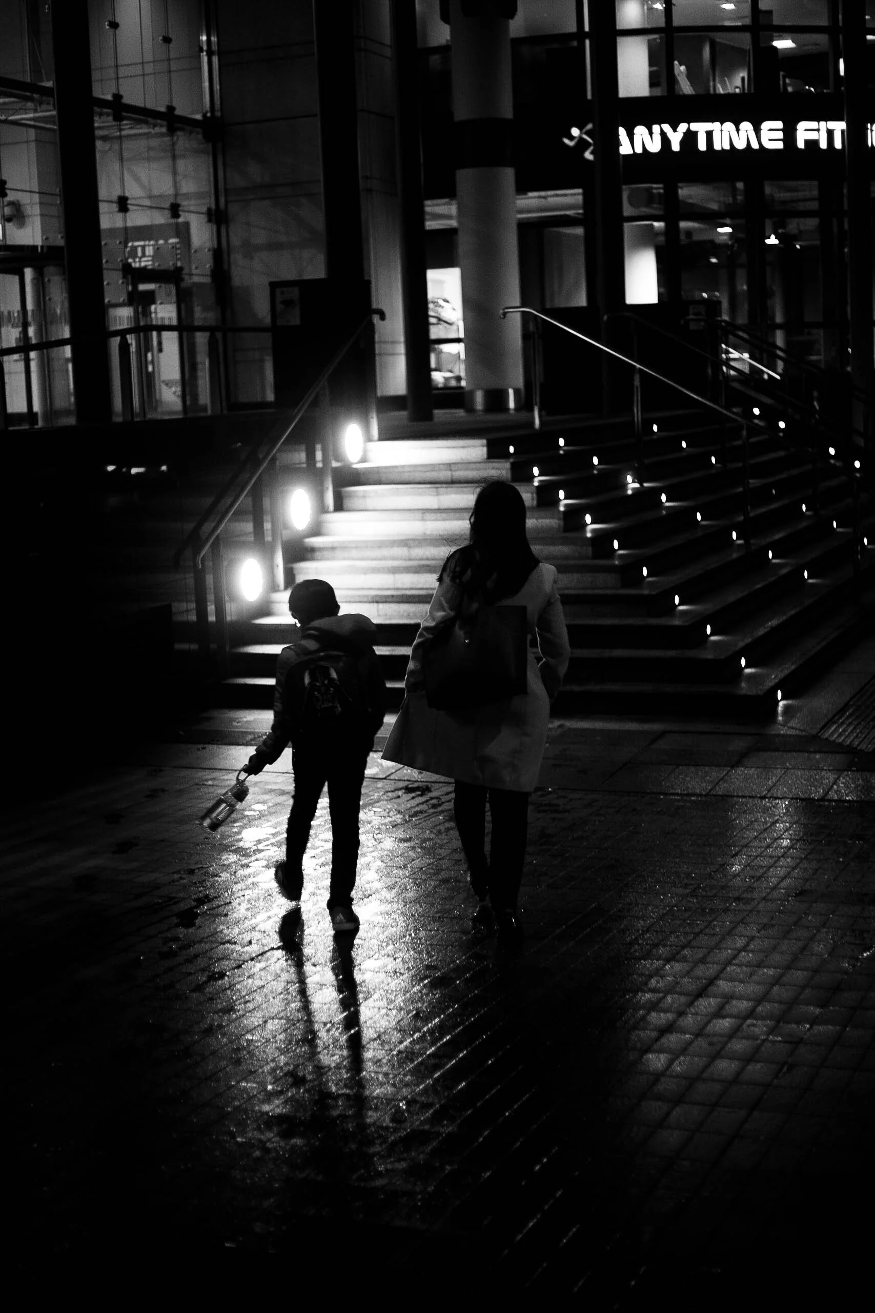 A silhouette of a woman and a child walking on a wet city street at night near steps with illuminated lights, and a sign that reads 'ANYTIME FIT' in the background.