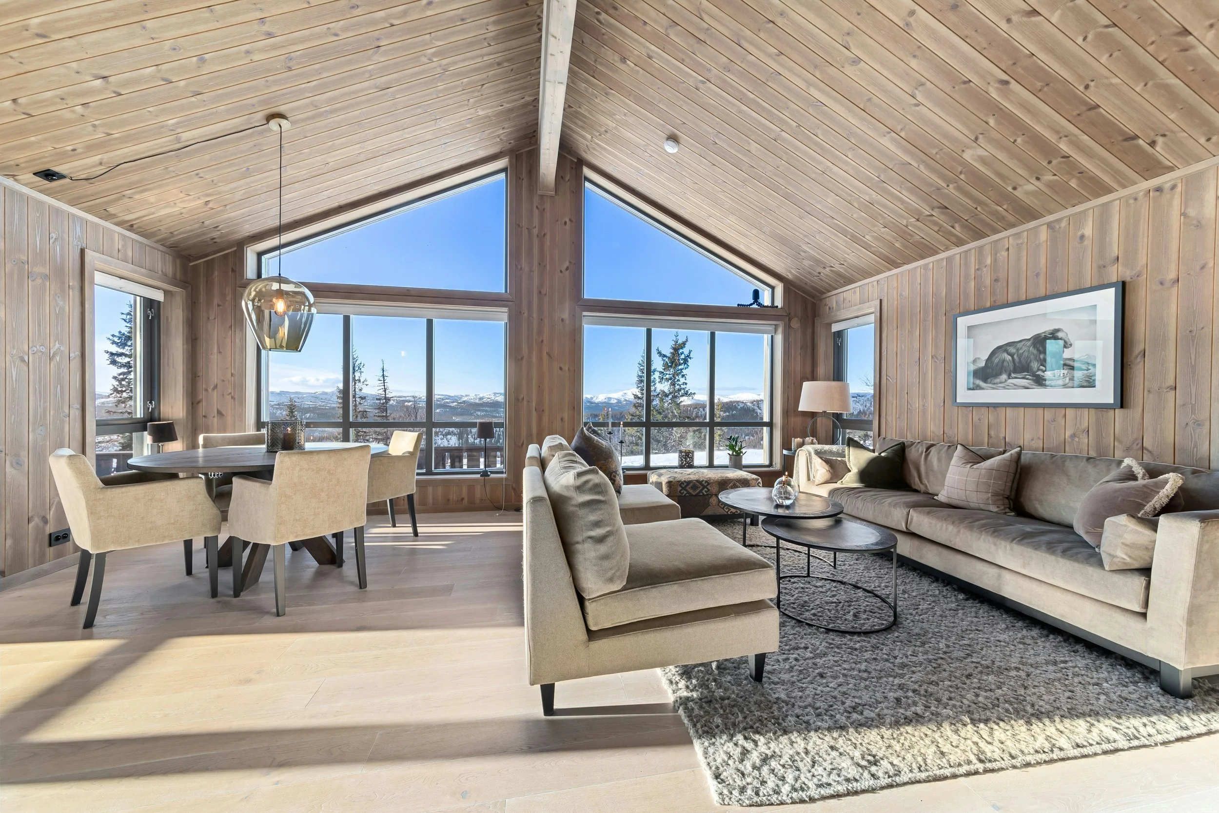 Living room with large windows showing mountain view, beige sectional sofa, round wooden coffee tables, dining area with six chairs, wood-paneled walls and ceiling, and artwork of a bear on the wall.