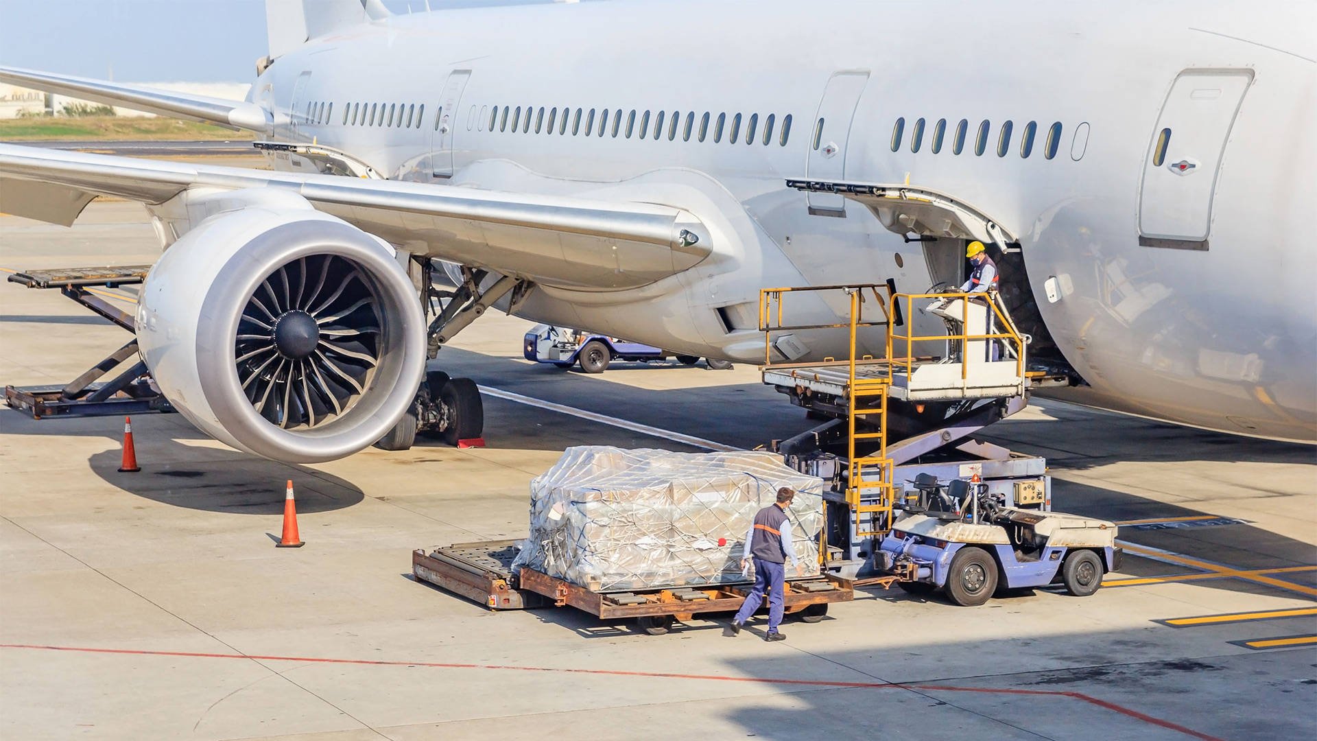 Airplane parked on tarmac with ground crew loading cargo. Technician on platform loading cargo into airplane's bay, with cargo wrapped and secured on pallet nearby.