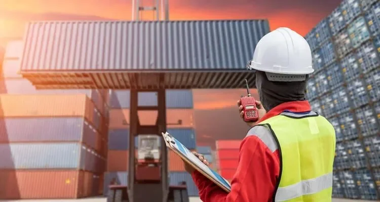 A worker in a safety vest and hard hat holding a walkie-talkie and clipboard at a shipping yard with stacked containers and a sunset sky.