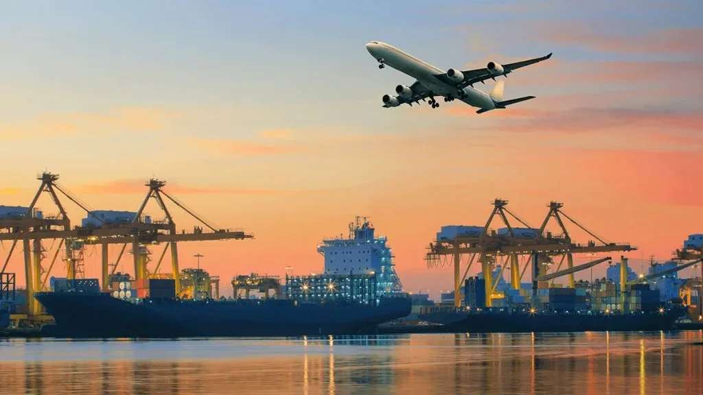A cargo port with ships and cranes at sunset, and an airplane flying overhead.