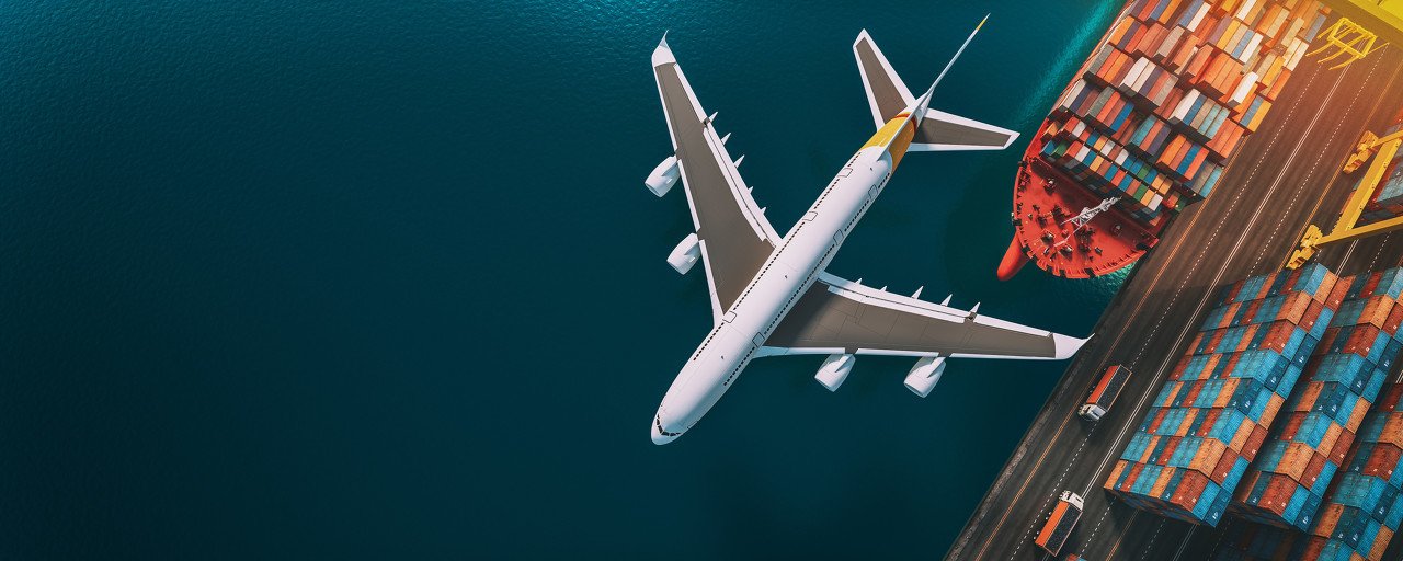 An airplane flying above a shipping port with cargo ships and containers.