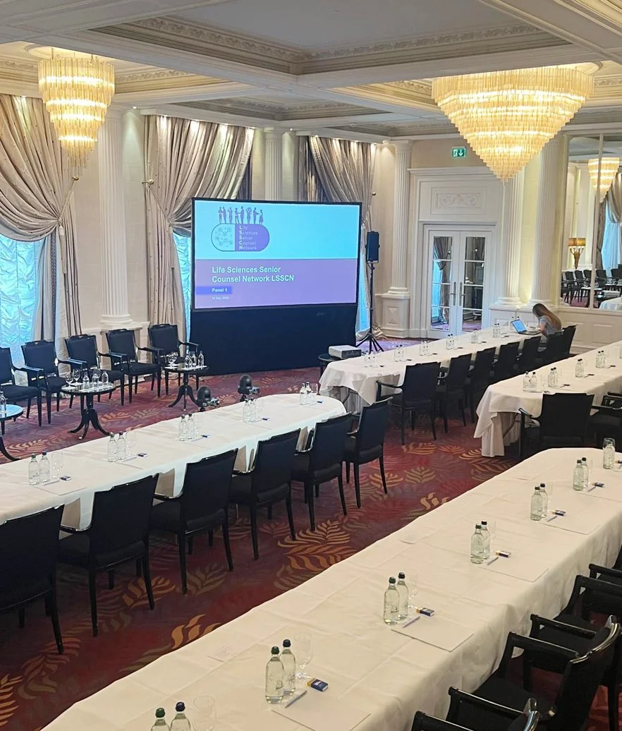 Konferenzraum mit länglichen Tischen, Stühlen, Wasserflaschen, Gläsern und einem großen Bildschirm, der eine Präsentation zeigt.