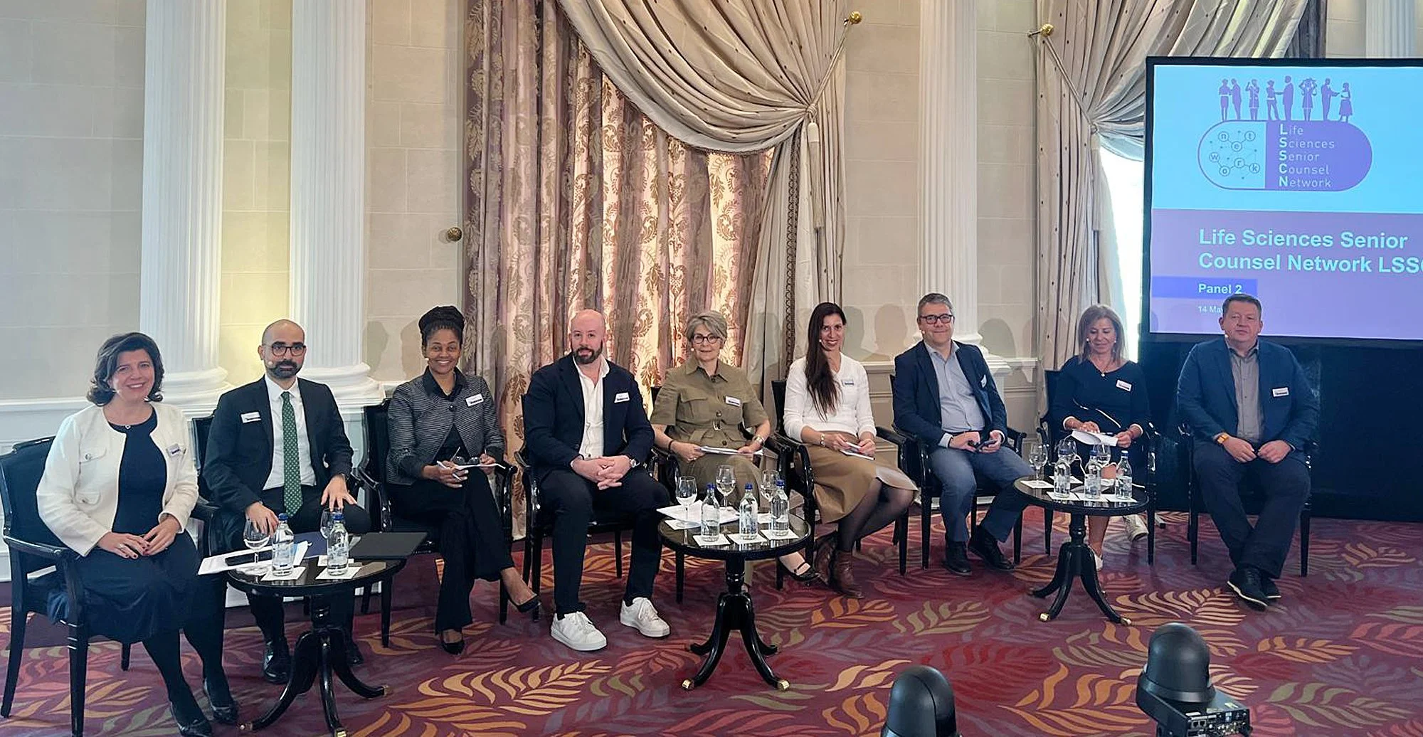 Neun Menschen sitzen in einem Panel bei einer Konferenz, mit einem Bildschirm auf der rechten Seite, der das Logo und den Titel der Veranstaltung zeigt, im Hintergrund sind große Vorhänge und Säulen.