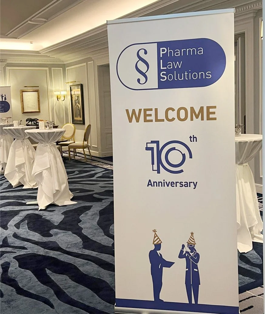 Ein Stehsäulenbanner mit dem Logo "Pharma Law Solutions" und dem Text "Willkommen zum 10. Jubiläum" bei einer Feier. Im Hintergrund sind Tische mit weißen Tischdecken und Stühlen zu sehen.