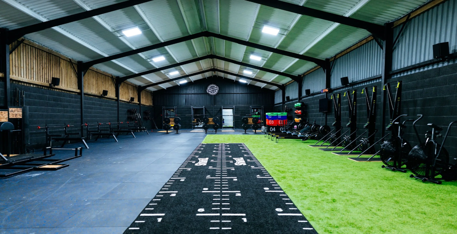 Interior of a modern gym with black walls and a high ceiling, featuring extensive facilities and equipment. Facilities cater to Olympic weightlifting, powerlifting, Hyrox, running, yoga, pilates, wellness, strength and conditioning, and gym classes.