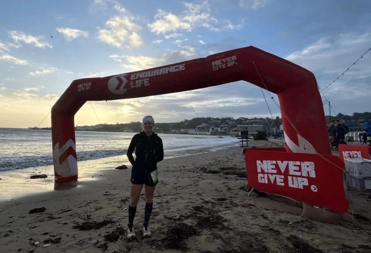 Person in athletic clothing standing on a beach under an arch with motivational slogans, with the ocean and a distant shoreline in the background during sunset.