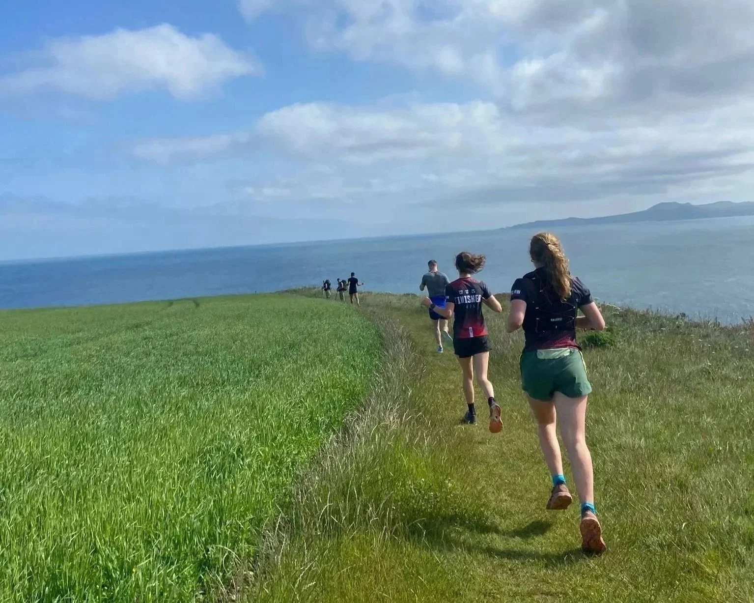 A group of people running along a narrow grassy trail beside a green field with the ocean in the background, under a partly cloudy sky.