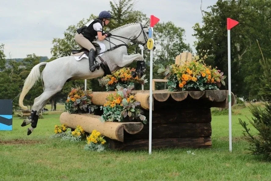 A rider and horse jumping over an obstacle decorated with flowers on a grassy field.