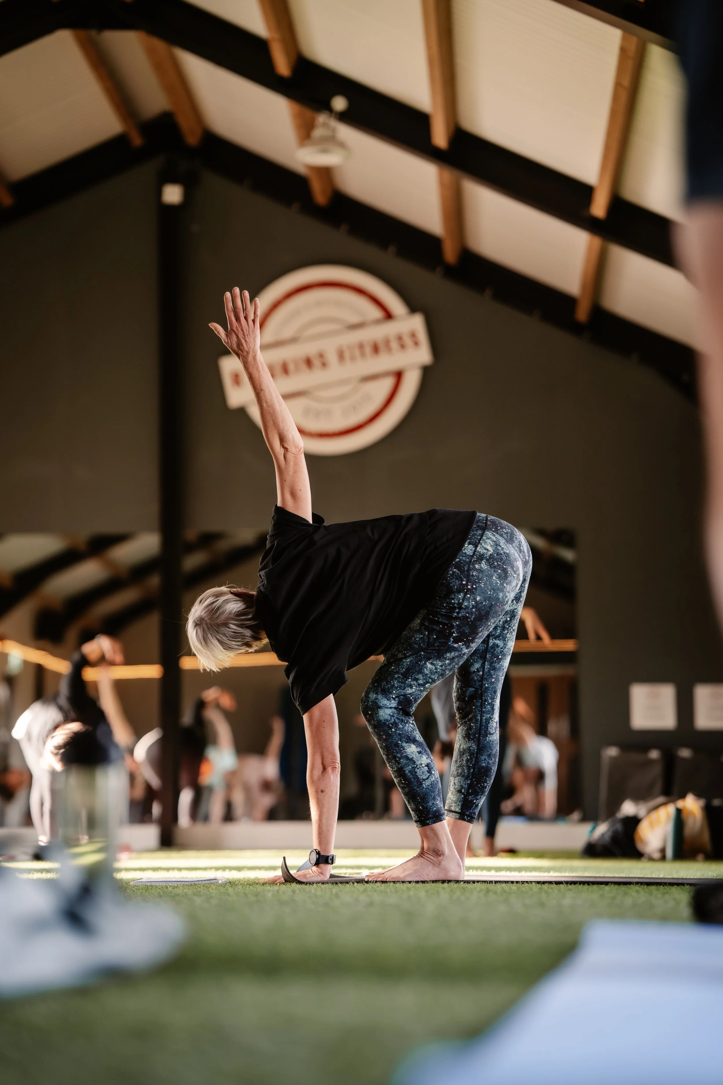 A woman practicing yoga in a studio, bending forward with one hand on the ground and the other arm raised, surrounded by other yoga students practicing.