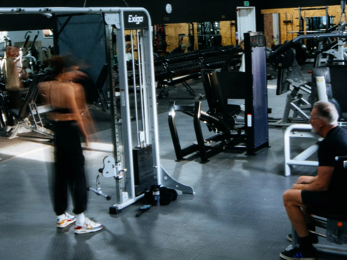 Two people working out in a gym with various exercise equipment, including a seated man in black athletic clothes and glasses, and a woman using a weight machine.