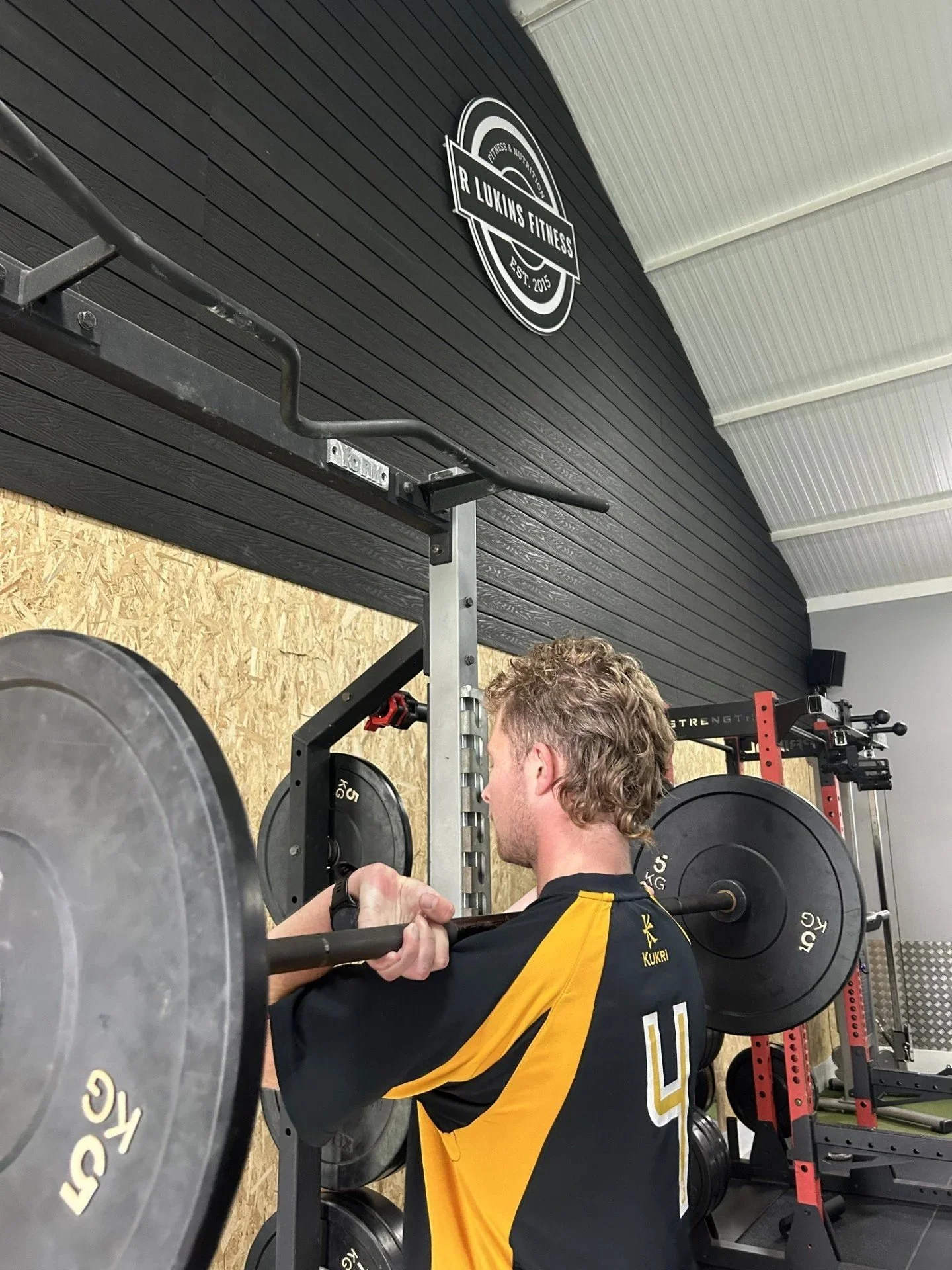 A person lifting a barbell with weights in a gym, facing a wall with a gym logo that reads 'R Lukins Fitness'.