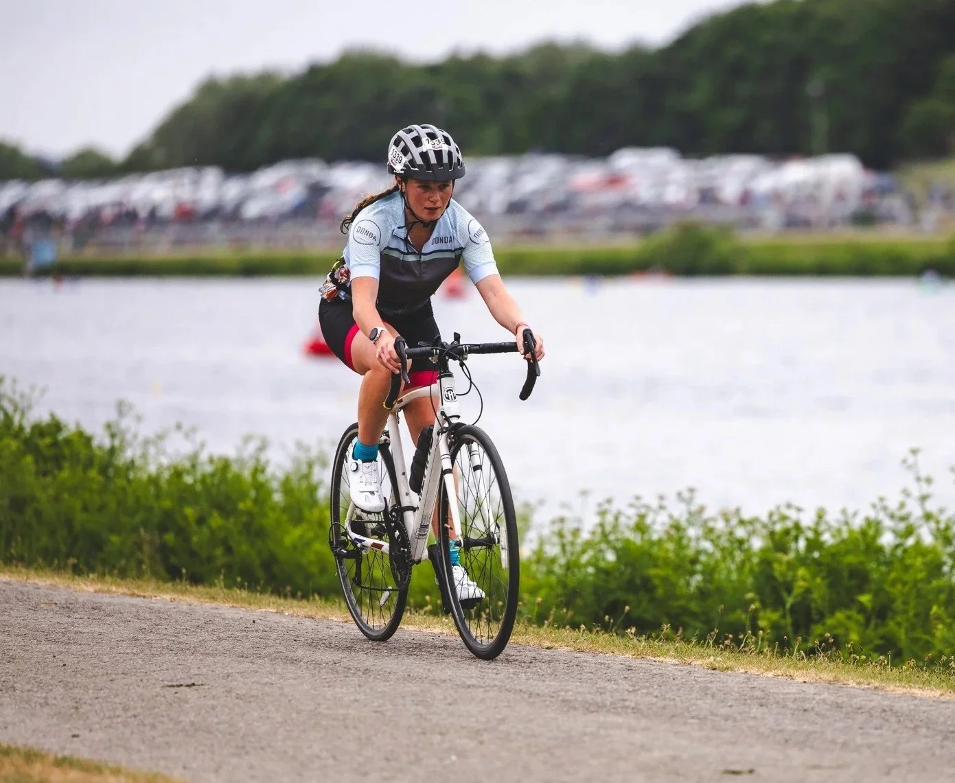 A woman riding a bicycle along a path next to a body of water, with a grassy area and a parking lot in the background.