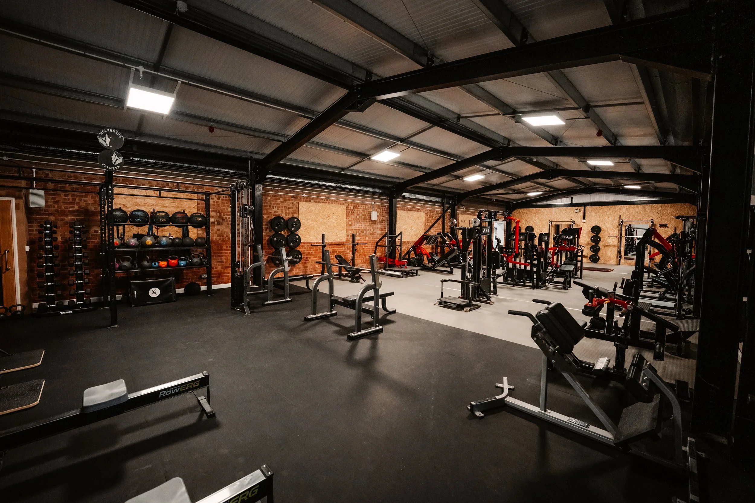 Interior view of a spacious gym with various workout equipment, including weight machines, free weights, battle ropes, and cardio machines, with brick walls and a high ceiling.