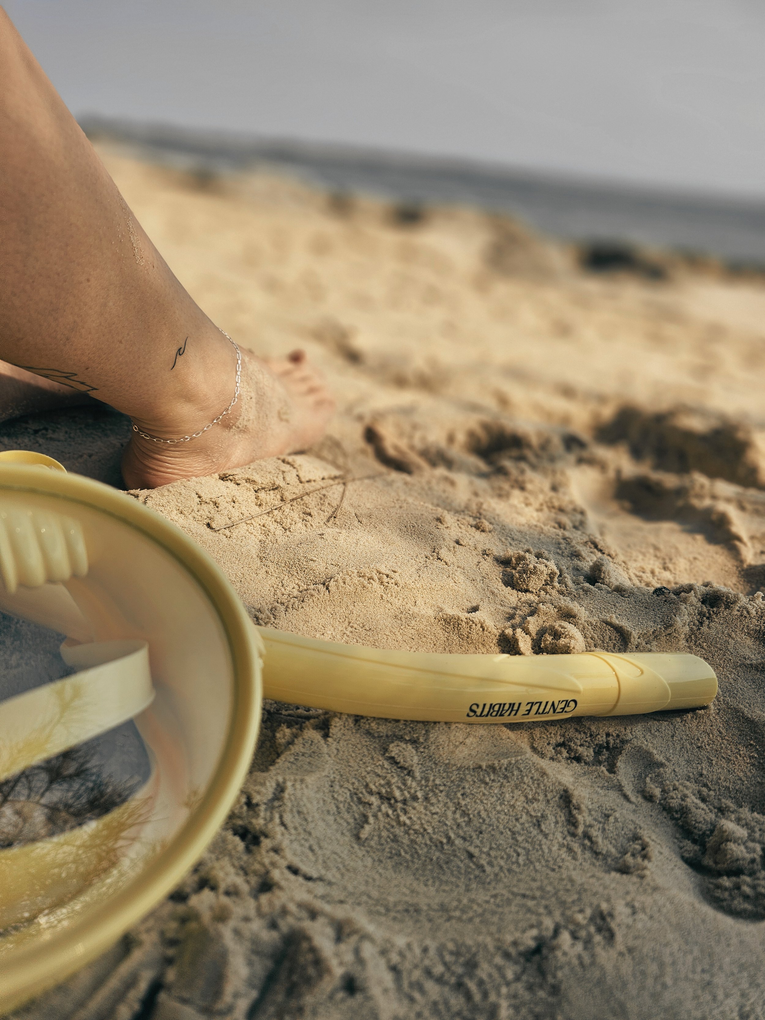 Close-up of a person relaxing on the beach with a yellow snorkel gear labeled 'GENTLE HABITS' resting on the sand, and the person's leg with an ankle bracelet in the background near the water.