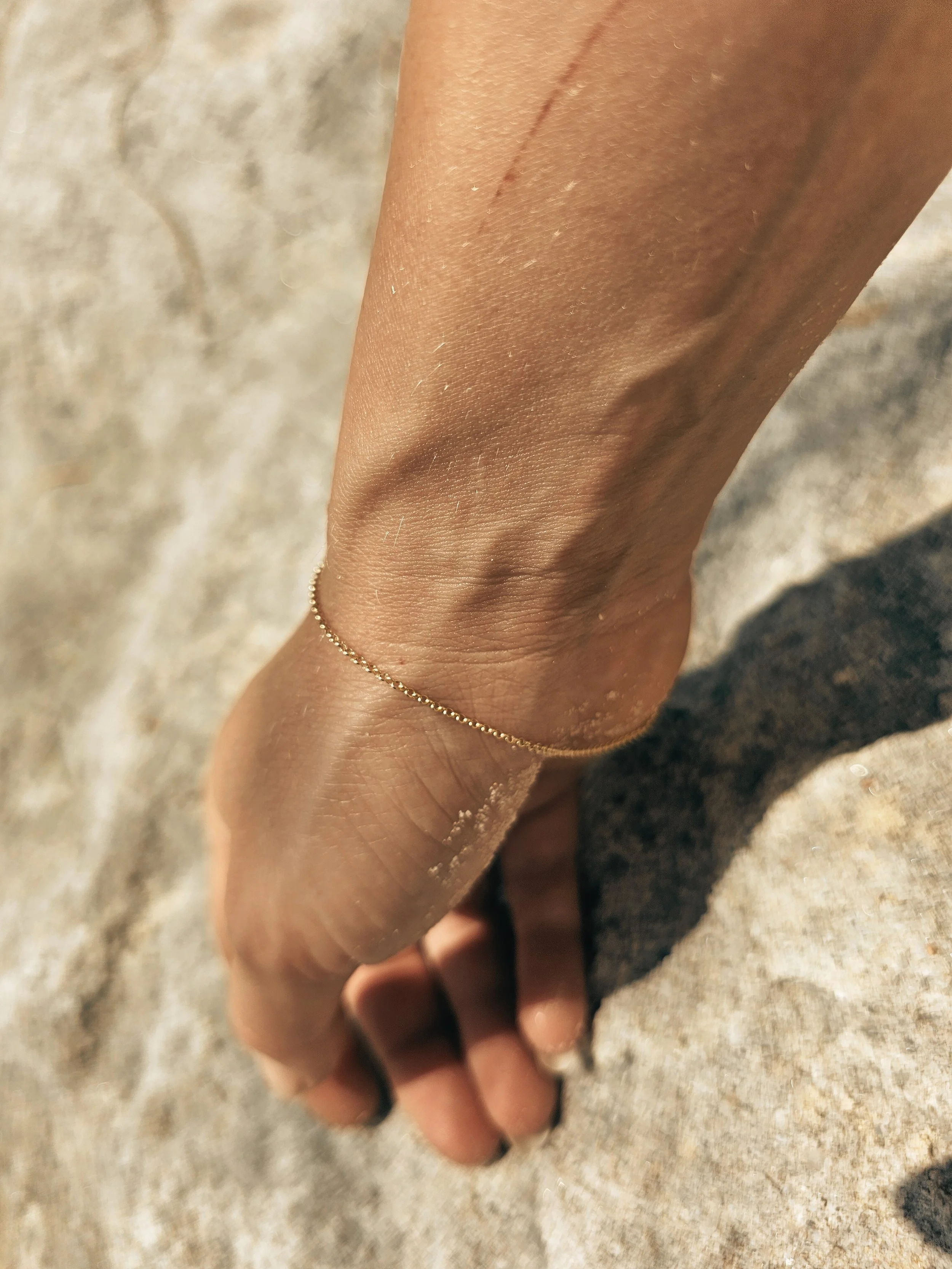 Close-up of a person's hand and wrist, wearing a delicate gold chain bracelet, against a sandy beach background.