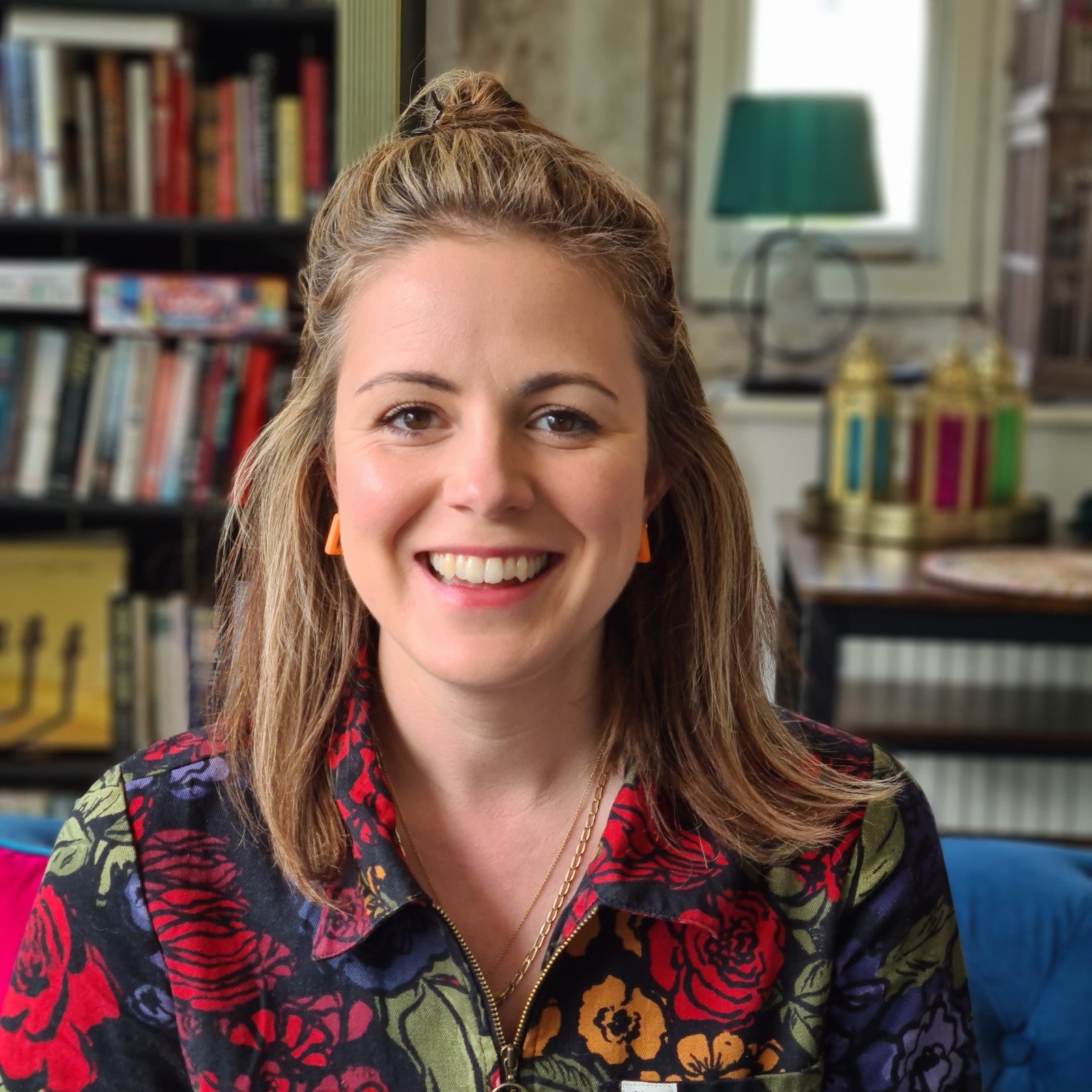 A smiling woman with light brown hair styled in a half-updo, wearing a colorful floral jacket, sitting in a room with bookshelves, a window, and decorative objects in the background.