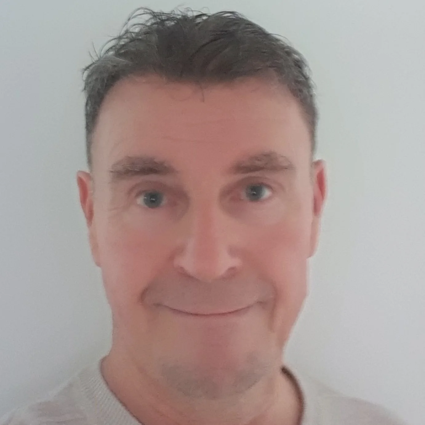 Close-up of a smiling man with short dark hair and blue eyes, wearing a light-colored shirt, against a plain light background.