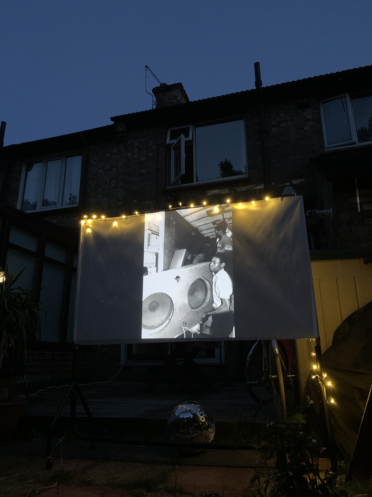 Projection d'une image en noir et blanc montrant deux hommes près de haut-parleurs, avec une maison en arrière-plan, lors d'une soirée en extérieur
