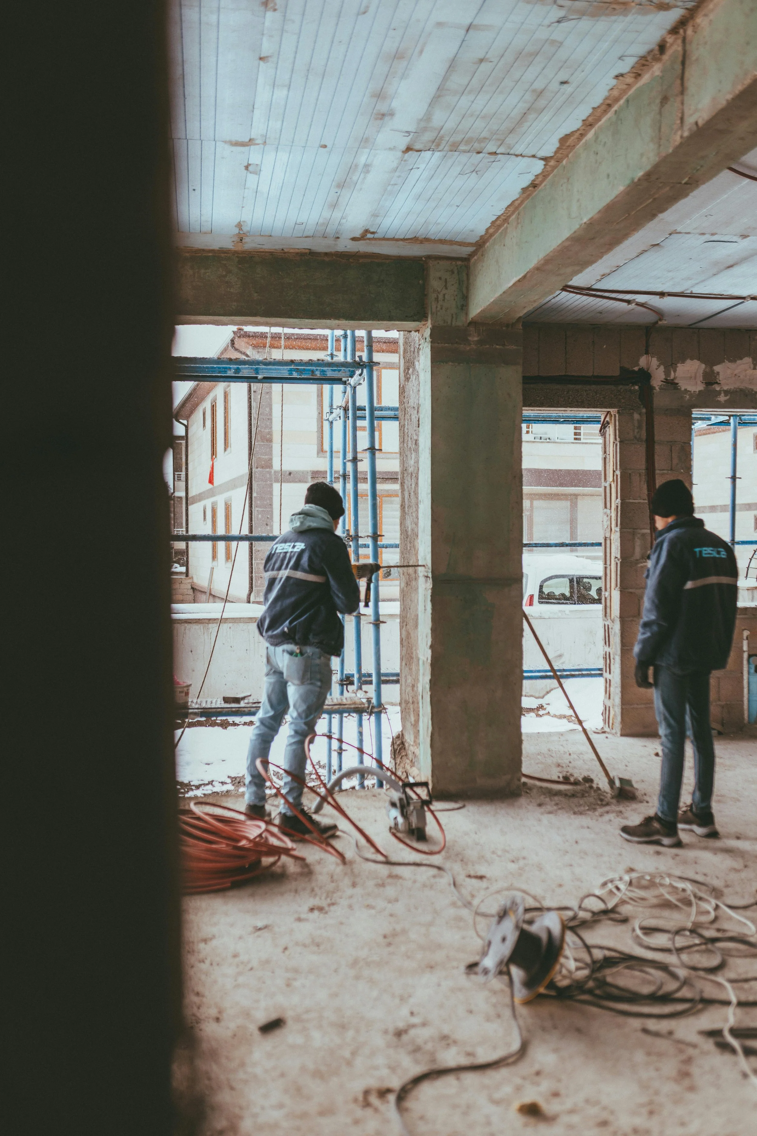 Construction workers in jackets working inside a building under construction, with scaffolding and construction materials on the unfinished floor.