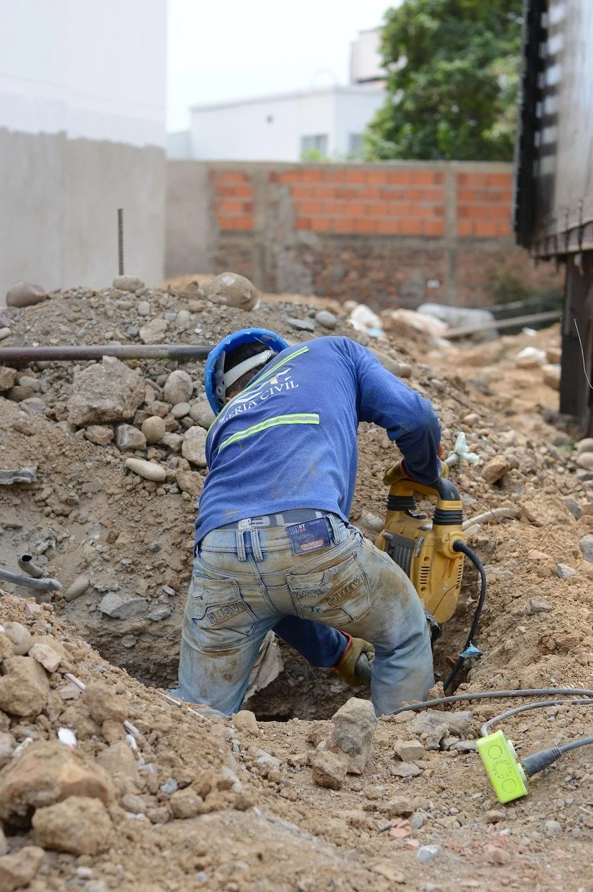 A construction worker wearing a blue helmet and a blue shirt working with a power tool in a dirt excavation site.