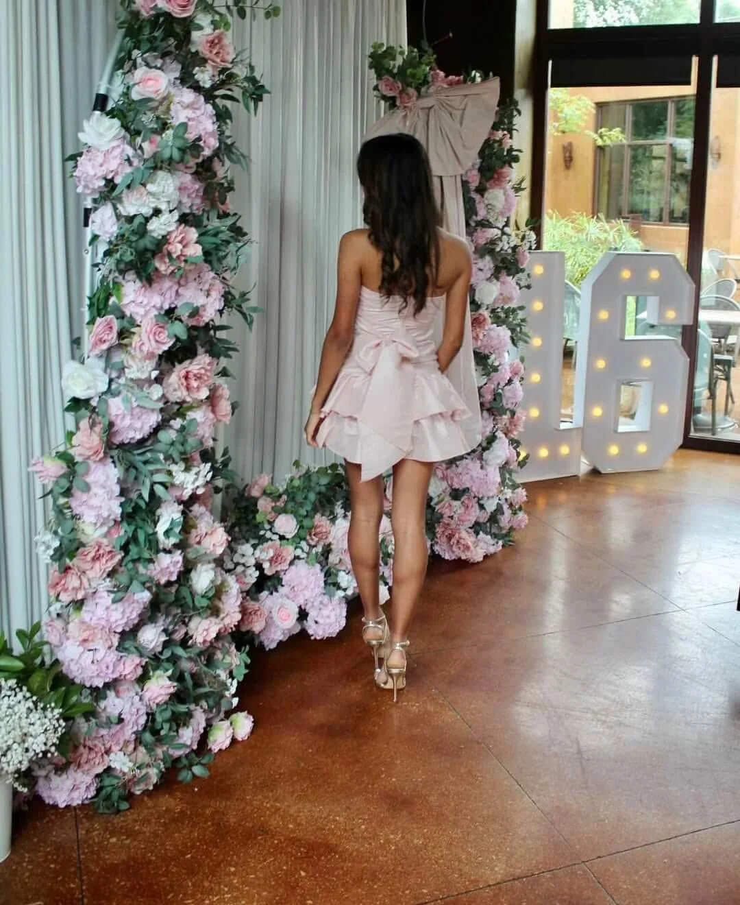 A woman in a pink dress and high heels standing in front of a floral arch with pink and white flowers, and a large lit-up 'LOVE' sign nearby, inside a room with large windows.