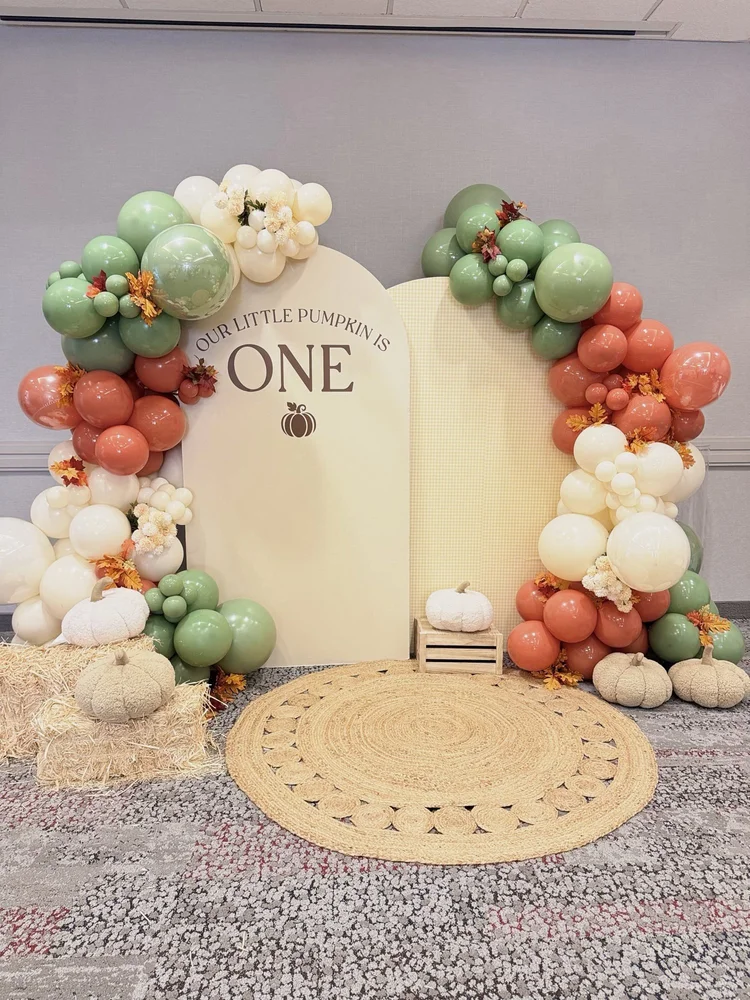 Fall pumpkin first birthday backdrop with arch panels, sage, terracotta and cream balloon garlands, plush pumpkins and a jute rug at Courtyard by Marriott Tysons McLean