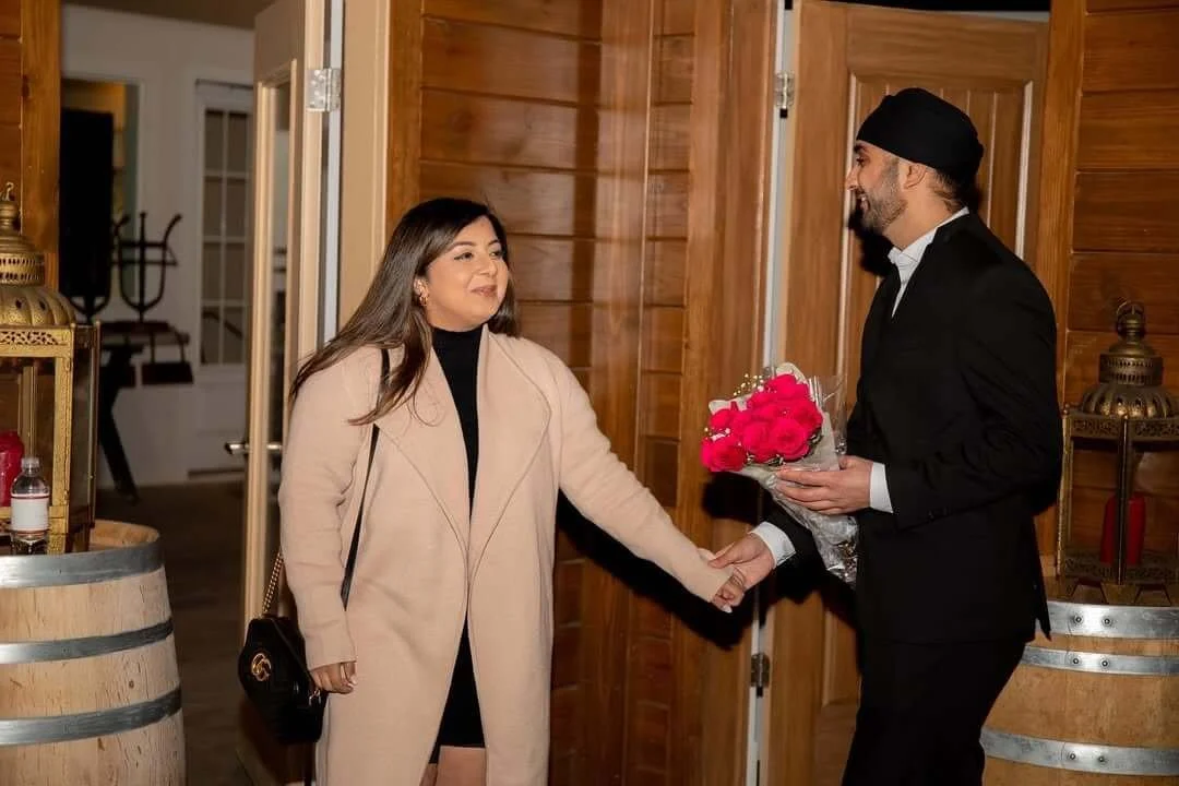 A woman is receiving a bouquet of pink roses from a man in a black suit and turban. They are shaking hands inside a warmly lit room with wood-paneled walls and décor.