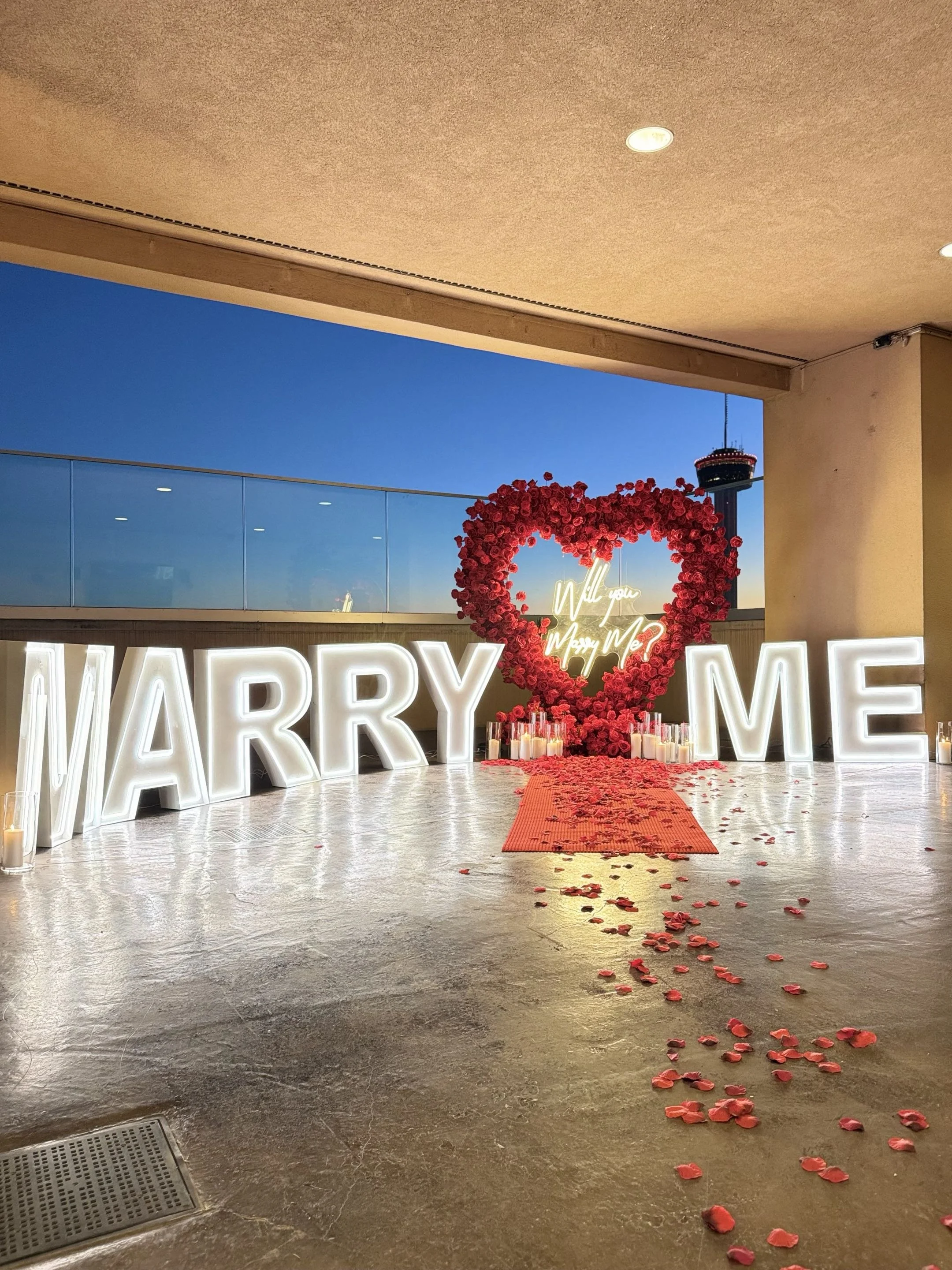 A decorative display at a wedding venue with large illuminated letters spelling 'MARRY ME,' a heart-shaped floral arrangement with red roses, a neon sign inside the heart that reads 'Will You Marry Me?', and a red rose petal aisle leading up to the display with some petals scattered on the floor.