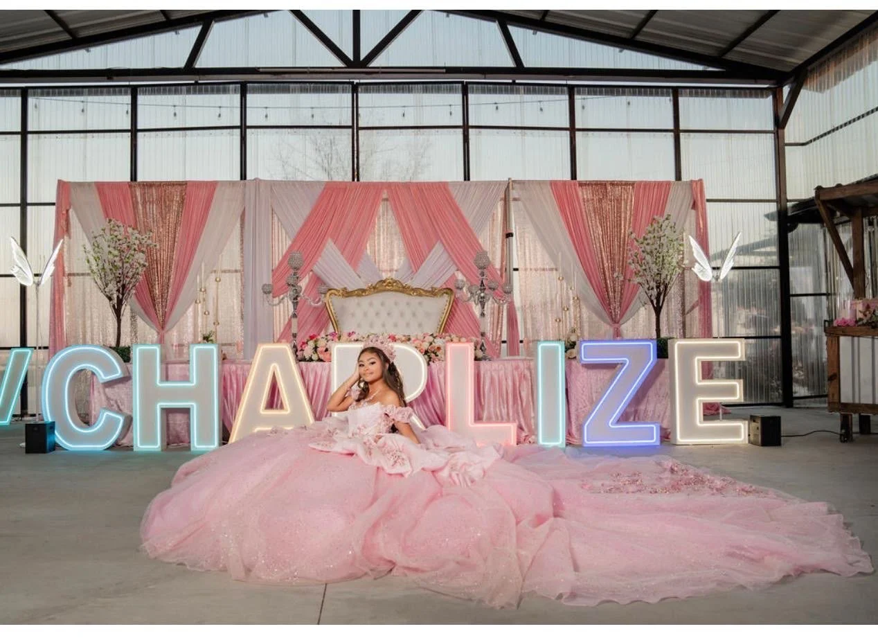 A young girl in a pink princess dress sitting on the floor in front of a decorated backdrop with large illuminated letters spelling 'CHAPIZ'. The backdrop features pink and white drapes, floral arrangements, and a white upholstered chair with gold accents. The setting appears to be inside a greenhouse or industrial space with large windows and metal framing.