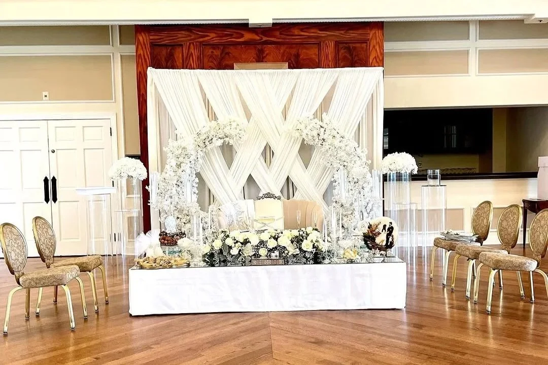 Elegant wedding reception setup featuring a backdrop with white draped fabric and floral decorations, a cushioned sofa, and chairs surrounding the display on a wooden floor.