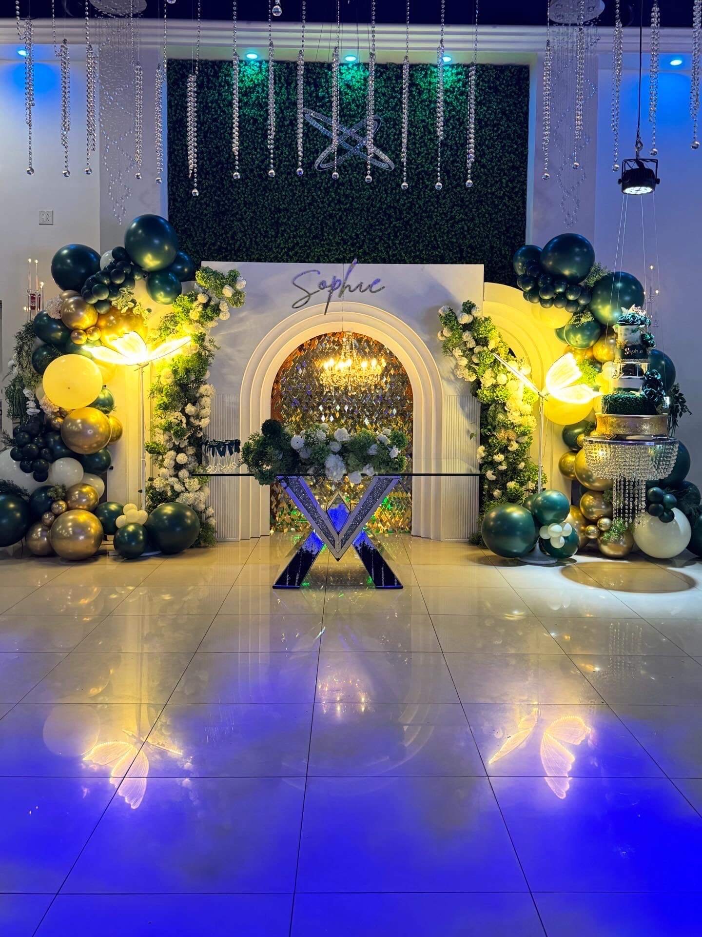 Elegant decorated stage with white arch, green and gold balloons, white flowers, and blue and yellow lighting, with a neon sign that reads 'Saphie' and a mirrored backdrop.