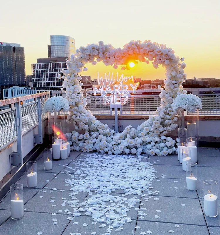 Sunset rooftop proposal setup with a white rose heart arch, Will You Marry Me neon sign, pillar candles and white petals at Mia Penthouse, the rooftop space at The Watermark Hotel in Tysons Corner, McLean, Virginia