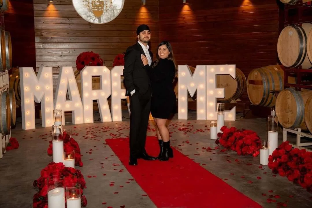 A couple standing on a red carpet in a wine cellar with large illuminated letters spelling 'MARRY ME' behind them, surrounded by red flowers and candles.
