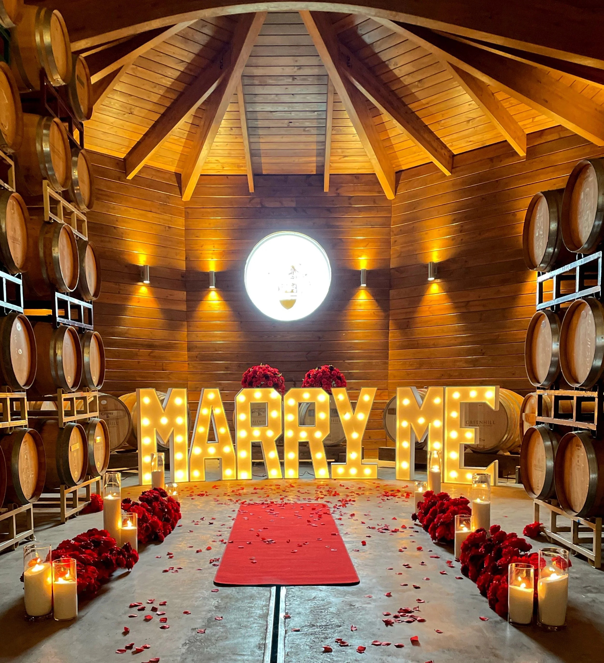 Wedding venue in a wine cellar with large illuminated letters spelling 'MARRY ME' at the end of a red carpet, surrounded by rose petals, candles, and floral arrangements, with wooden wine barrels on shelves and warm lighting.