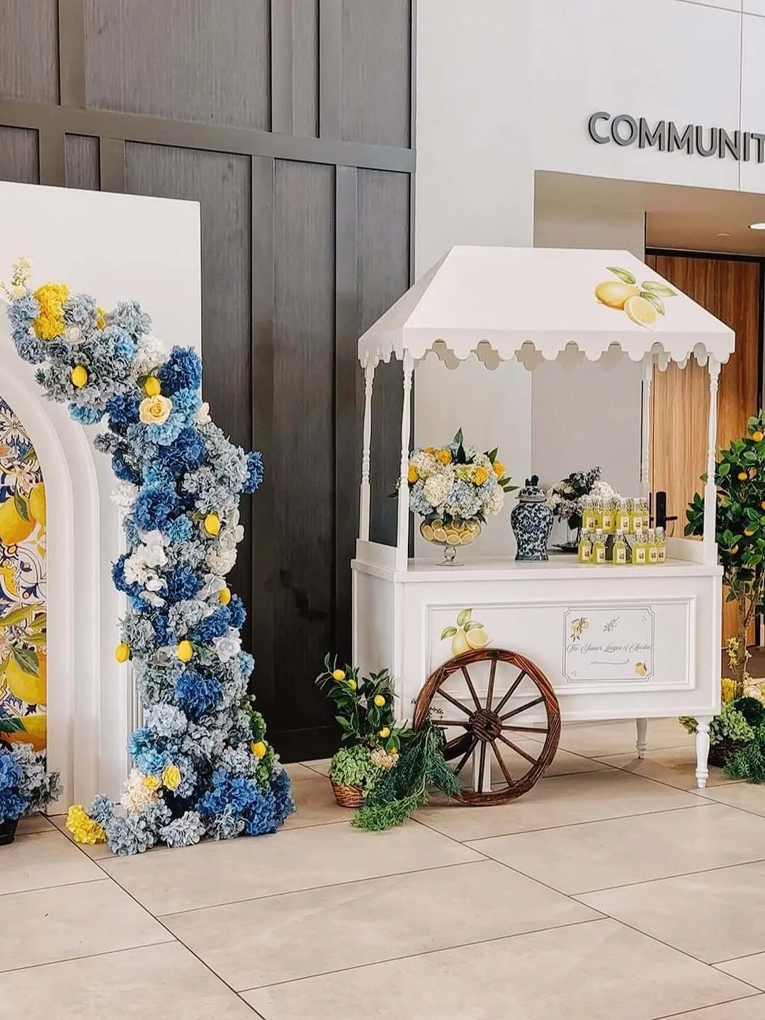 A white cart with a scalloped canopy, decorated with lemon illustrations, surrounded by floral arrangements and greenery, near a dark wooden wall and a sign that reads 'COMMUNITY'.