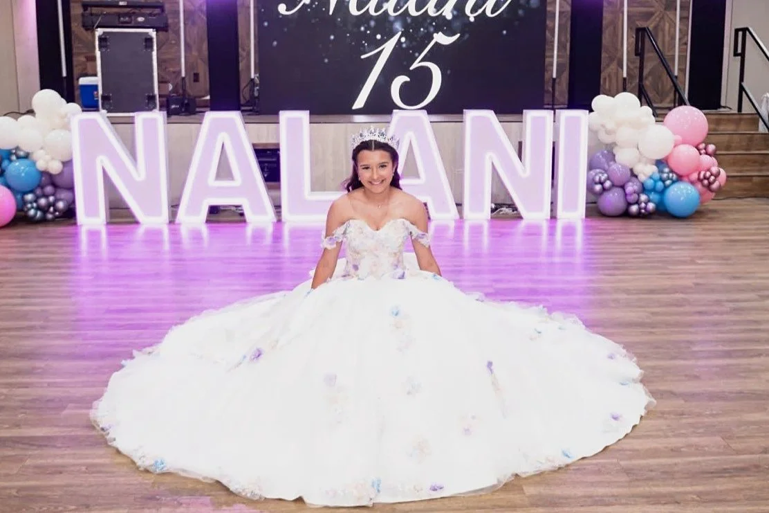 Quinceañera honoree in a white floral ball gown in front of lavender lit marquee letters with a 15 sign and pastel pink, blue and lilac balloon garlands