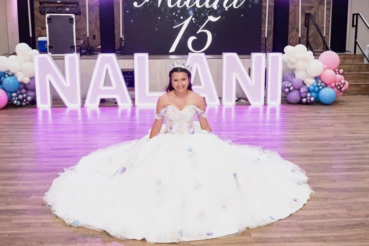 Quinceañera honoree in a white floral ball gown in front of lavender lit marquee letters with a 15 sign and pastel pink, blue and lilac balloon garlands