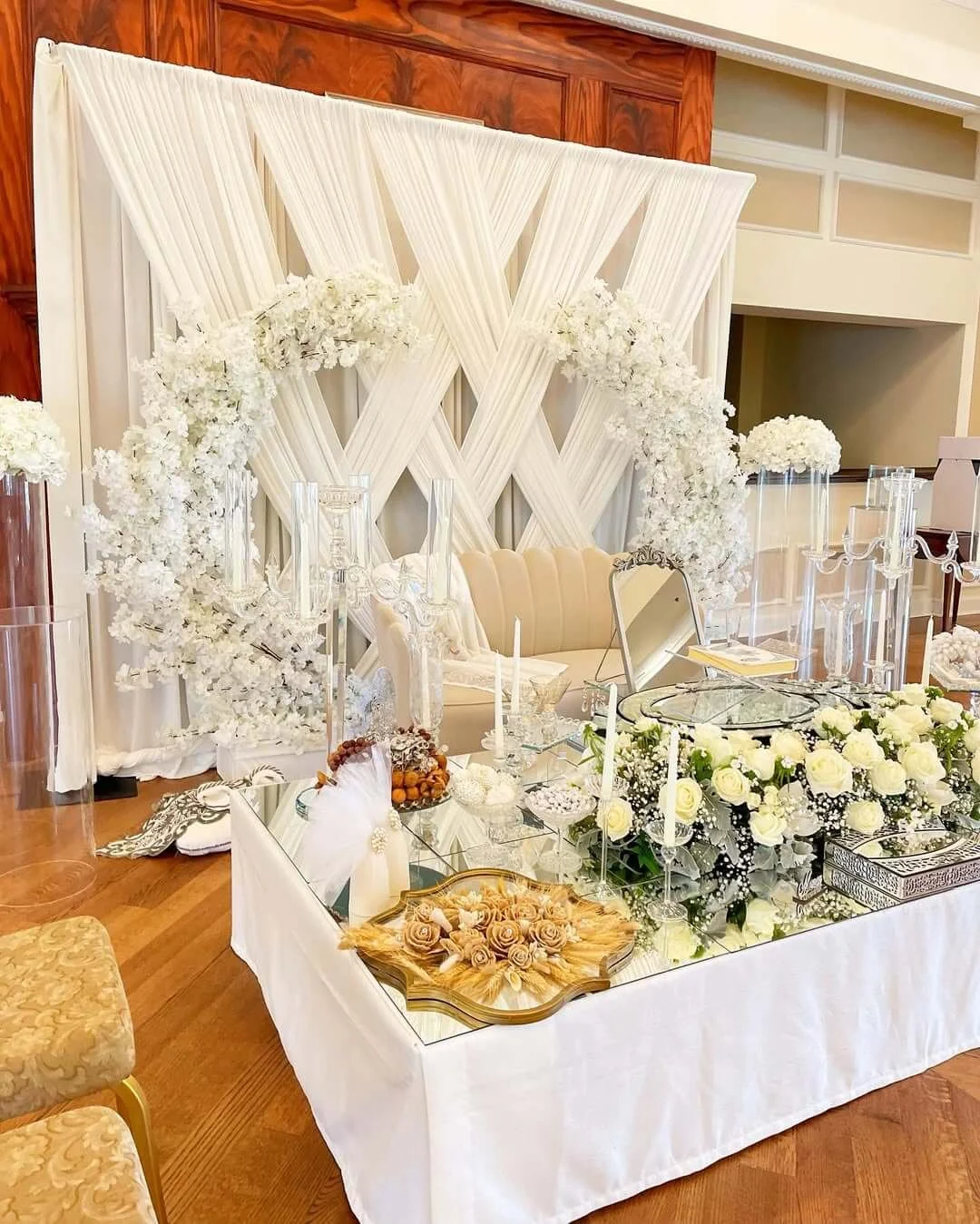 Elegant wedding or event setup with a white floral backdrop, white floral arrangements on the table, candles, and mirror decorations.