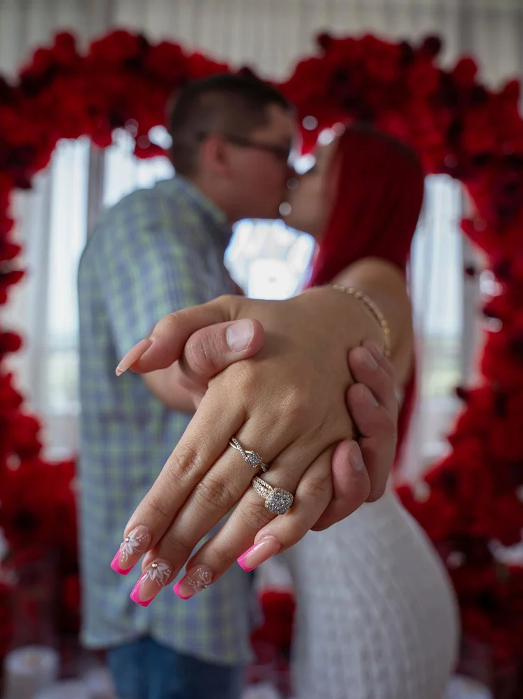Newly engaged couple showing their engagement rings in front of a red rose heart arch in McLean, Virginia