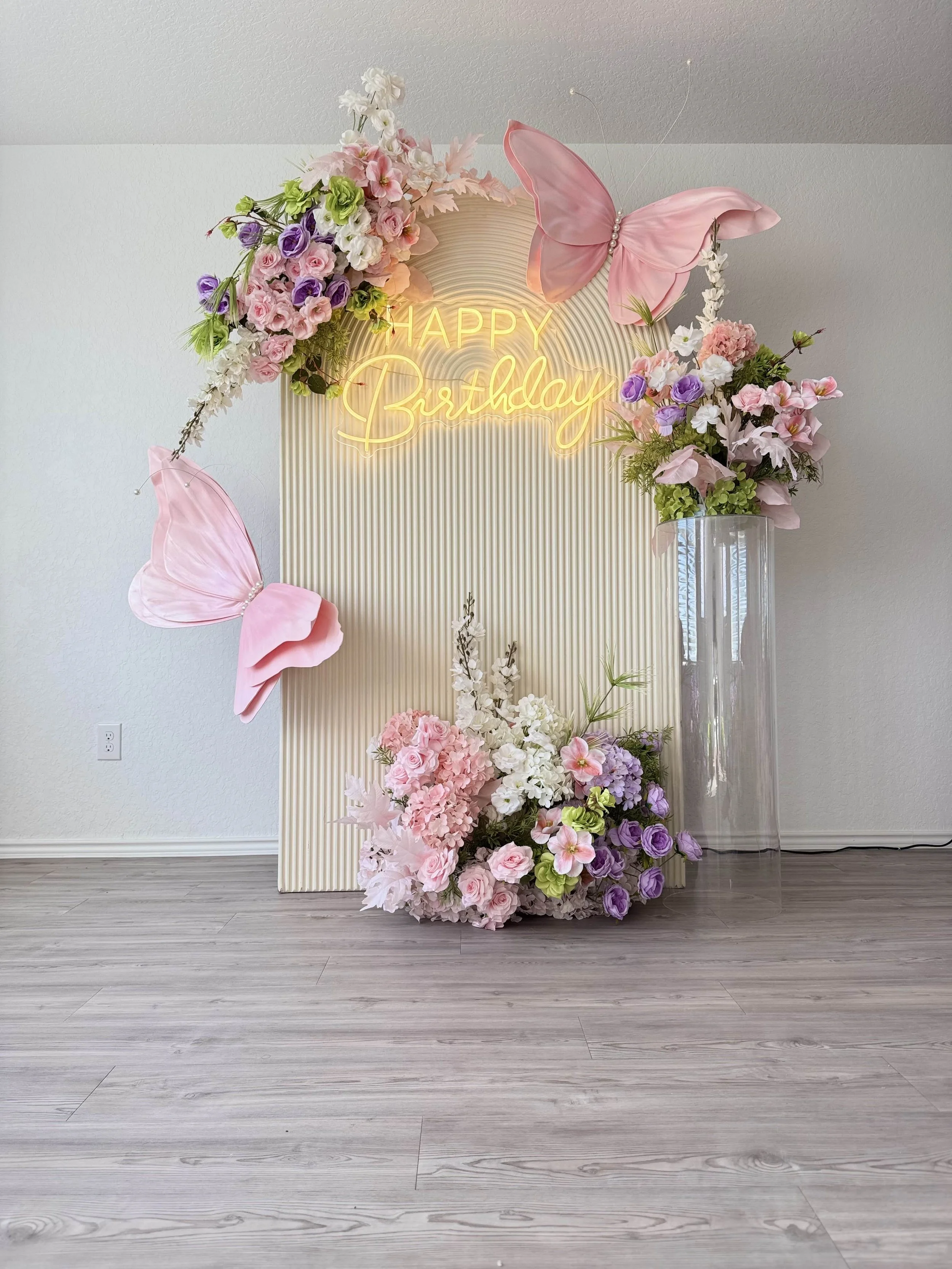 Decorative birthday display with pink and purple flowers, a neon sign saying 'Happy Birthday,' pink butterfly decorations, and a tall glass vase on a white ribbed backdrop on a wooden floor.