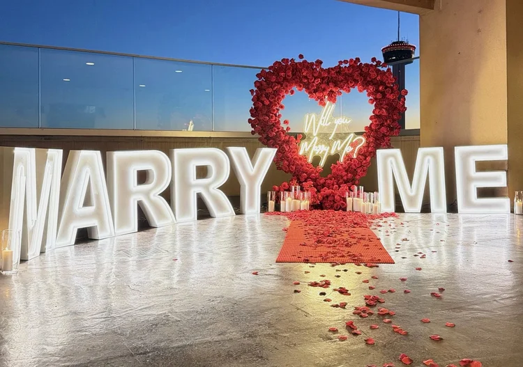 Large lit MARRY ME letters with a red rose heart arch, neon sign, pillar candles and a red carpet with rose petals