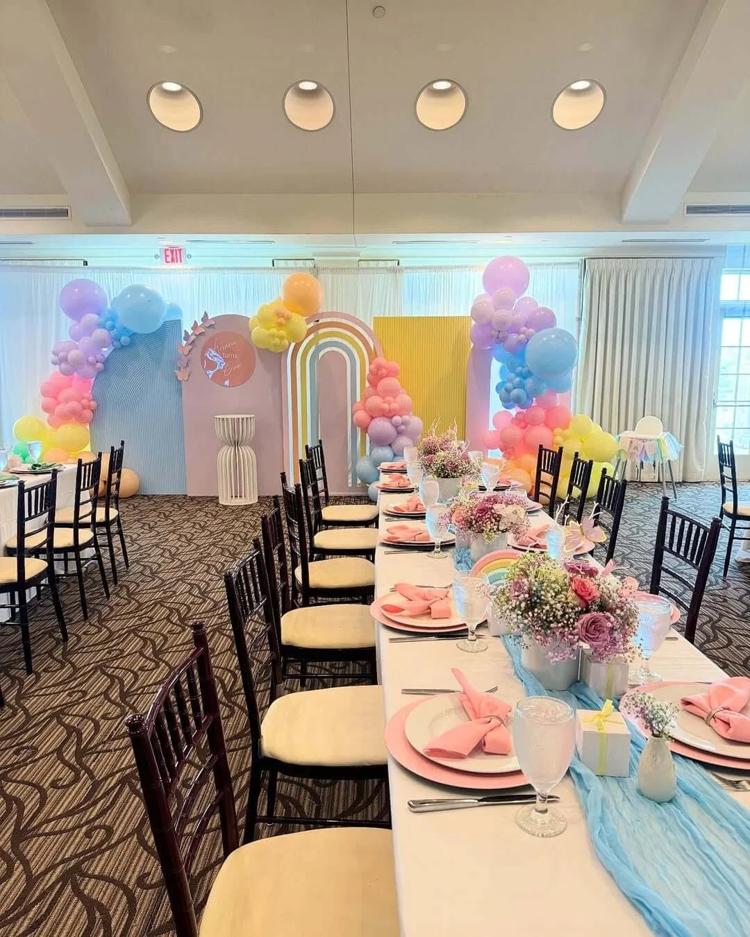 Decorated banquet table with pink napkins, floral centerpieces, and pastel balloons in front of a rainbow-themed backdrop for a celebration.